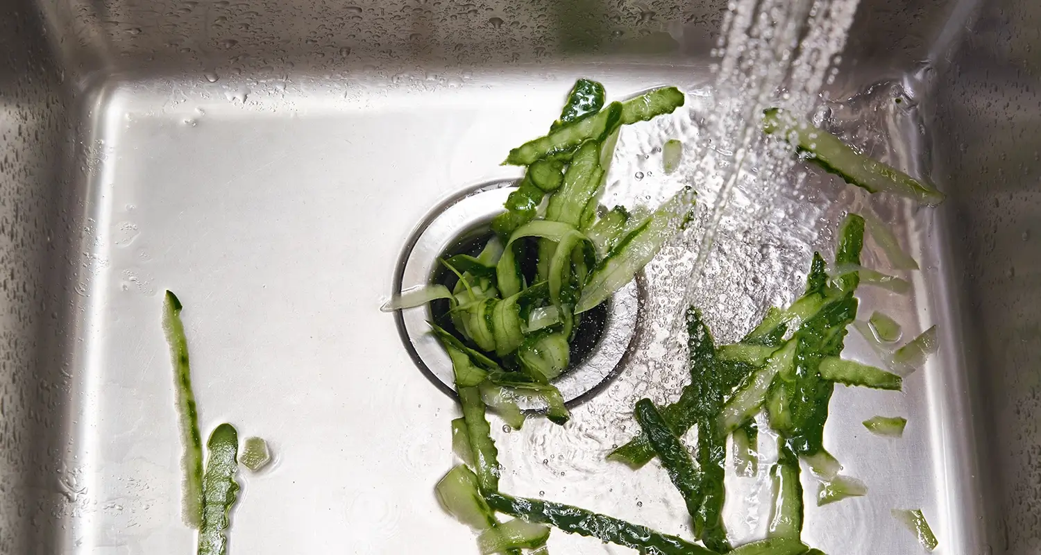 Water spraying over the peels of a cucumber in the bottom of a kitchen sink with a garbage disposal.